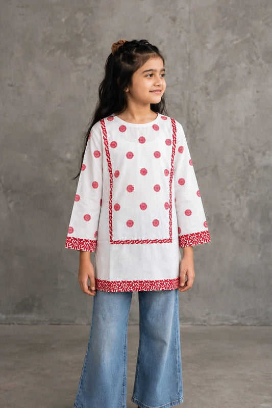 A young girl wearing a white three-quarter sleeve kurta with red polka dots and red patterned trim, paired with light blue jeans, standing against a textured gray studio background.